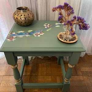 May include: A sage green side table with a hand-painted floral design. The table has gold accents on the legs and holds a gold lantern and a small vase of purple flowers. The table is approximately 61 cm tall.