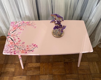 May include: A light pink rectangular table with rounded corners and four legs. The table's top features a floral design of pink cherry blossoms on a branch. A small glass vase with purple flowers sits on the table.