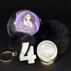 May include: A black and silver glitter keychain with a large silver number 40, a small round container of glitter, and a clear plastic sphere with a cartoon image of a woman with a face mask and the text "The Glitter Doctor".