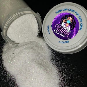 May include: A clear plastic container tipped over, spilling fine white iridescent glitter onto a black surface. The white lid features a purple circular label with the text "The Glitter Doctor" and an illustration of a medical professional.