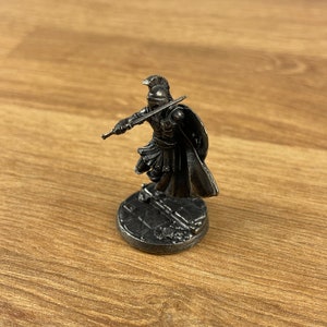 May include: A small, silver figurine of a warrior in a helmet and armour, holding a sword and shield. The figurine stands on a circular base with a textured design. The warrior is in a dynamic pose, as if in battle.
