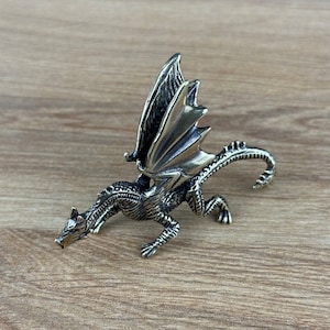 May include: A silver-coloured dragon figurine with outstretched wings, perched on a wooden surface. The dragon's head is lowered, and its tail curls around its body.