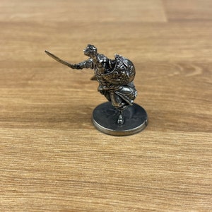 May include: A silver miniature figurine of a warrior holding a sword and shield. The warrior is wearing armour and a helmet. The figurine is standing on a round base.