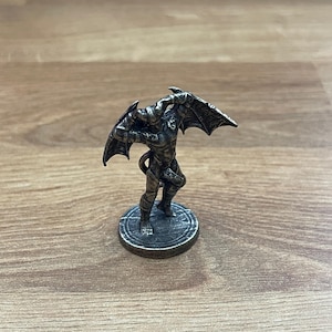 Op de afbeelding: Een zilverkleurig miniatuurbeeldje van een demon met vleugels, staande op een ronde basis. De demon houdt zijn vleugels vast met zijn handen en heeft een staart.