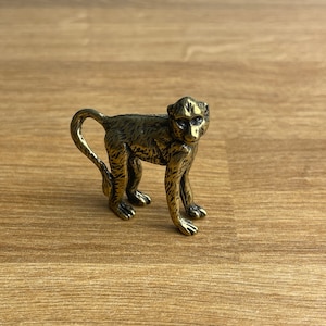 Può includere: Una piccola statuetta di scimmia dorata con una superficie testurizzata. La scimmia è in piedi su tutti e quattro gli arti con la coda arrotolata attorno al suo corpo.