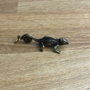 May include: A small, detailed, bronze chameleon figurine with a curled tail. The figurine is on a wooden surface.