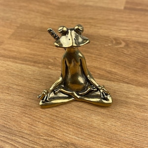 May include: A gold-coloured frog figurine sitting in a meditative pose with a small sword behind its head.