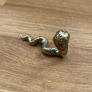 Könnte beinhalten: Eine kleine, goldfarbene Metallfigur einer Kobra-Schlange mit erhobenem Kopf und ausgebreiteter Haube. Die Schlange ist in einer Ruheposition zusammengerollt.