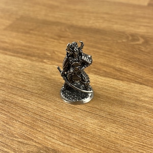 May include: A detailed miniature samurai warrior figurine, crafted from a metallic material. The figure is posed with a katana and shield, showcasing intricate armor details. The base is circular, and the overall design is a dark, antique silver color.