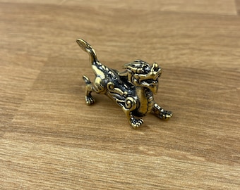 Puede incluir: Una pequeña figura de metal dorada de un dragón con detalles intrincados. El dragón está de pie sobre sus cuatro patas con la cabeza levantada y la boca abierta.