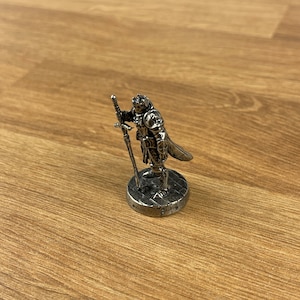 May include: A silver miniature figurine of a knight in armour holding a sword. The knight is standing on a round base with a textured surface.