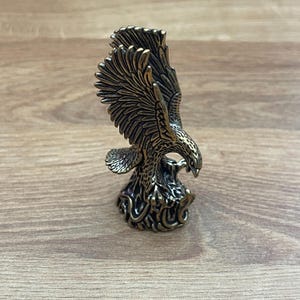 Könnte beinhalten: Eine goldfarbene Adlerfigur aus Metall mit ausgebreiteten Flügeln, die auf einem Sockel mit Schlangenmuster sitzt.