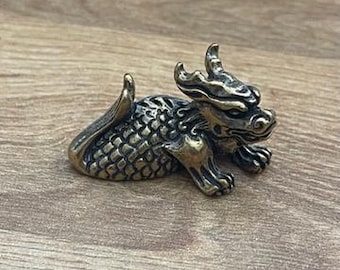 Puede incluir: Una pequeña figura de dragón de color dorado con detalles intrincados. El dragón está enroscado en una posición de descanso con la cabeza girada hacia un lado. La figura está hecha de metal y tiene una superficie texturizada.