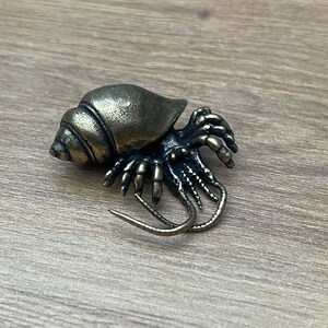 May include: A bronze figurine of a hermit crab with detailed legs and a shell. The figurine is on a wooden surface.