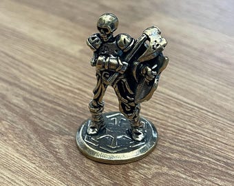 Puede incluir: Una figurita de metal en miniatura de un guerrero esqueleto sosteniendo un hacha y un escudo. La figurita está de pie sobre una base redonda con un diseño decorativo.