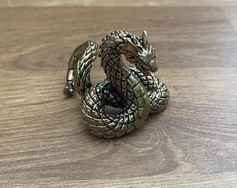 May include: A gold-colored metal dragon figurine curled up in a coiled position. The dragon has detailed scales and a pointed snout.