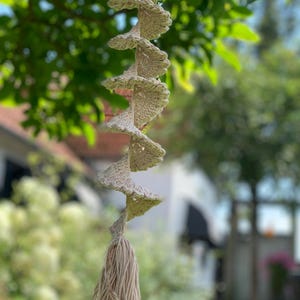 May include: A beige macrame hanging with a tassel at the bottom. The hanging is made of a series of crocheted loops that are twisted together to form a spiral shape.