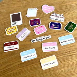 Op de afbeelding: Een verzameling kleurrijke stickers met verschillende zinnen en ontwerpen. De stickers zijn gedrukt op wit papier en hebben een glanzende afwerking. Sommige van de zinnen zijn "Hello I'm delusional", "Hello I'm avoiding reality", "Who did that to you?", "Read all night book club", "Sci-fi book club", "Romance book club", "Smut book club", "Fantasy book club", "Either give me more faery wine or leave me alone", "Fictional or delusional er", "Pantone Morally Grey", "Hello I'm taking theoretical exams", "Hello I'm busy reading", "Hello I'm avoiding talking to you", "Hello I'm probably reading smut".