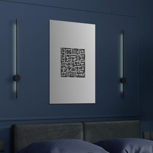 Op de afbeelding: Een zwart-wit kalligrafieprint van Arabische schrift in een vierkant frame, hangend aan een donkerblauwe muur boven een bed met een zwart hoofdeinde.