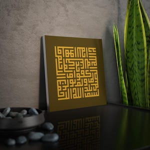 Op de afbeelding: Een vierkante canvas print met gouden Arabische kalligrafie op een donker olijfgroene achtergrond. Het kunstwerk wordt getoond op een zwart oppervlak, met een donkergrijze muur op de achtergrond. Een decoratieve kom met zwarte stenen en een groene plant zijn ook zichtbaar.