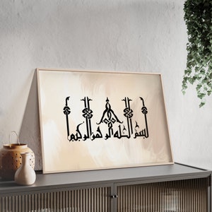 Op de afbeelding: Ingelijst kunstwerk met zwarte Arabische kalligrafie op een abstracte beige en witte achtergrond. De kalligrafie is gecentreerd en omlijst in een licht houten frame. Het kunstwerk wordt tentoongesteld op een donkergrijze kast.