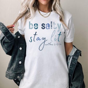 以下が含まれることがあります： 白いTシャツに、青と白のグラフィックがプリントされています。グラフィックには「be salty stay lit」と「matthew 5:13-14」の文字が書かれています。