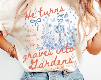 May include: Ivory-colored sweatshirt featuring a skeleton illustration surrounded by blue flowers and butterflies. The text reads "He turns dry bones come to life graves into Gardens" with "Ezekiel 37" below. The design includes a bow and star accents.