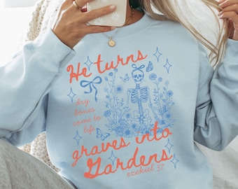 May include: Light blue sweatshirt with the text "He turns dry bones come to life graves into Gardens" and a skeleton graphic surrounded by flowers and butterflies. The text is in coral and blue.