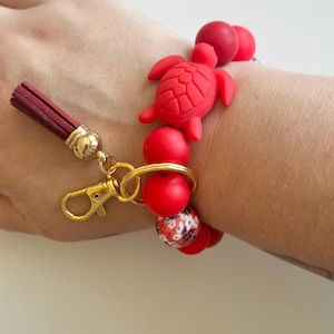 May include: Red beaded bracelet with a red turtle charm and a tassel. The bracelet has a gold-coloured clasp and a mix of solid red and patterned beads. The tassel is burgundy with a gold-coloured cap.