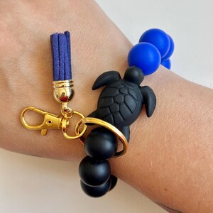 May include: A black turtle keyring with a gold clasp, a navy blue tassel, and black and blue beads. The keyring is attached to a gold ring. The beads are round and smooth.