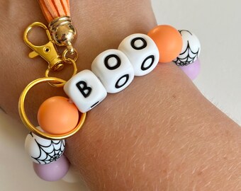 Llavero elástico con cuentas de silicona en tonos naranja pastel y morado con telaraña y letras.