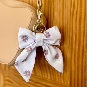 Könnte beinhalten: Ein helllila Stoff-Schleifen-Schlüsselanhänger mit einem Blumenmuster aus lila Blumen und grünen Stielen. Die Schleife ist an einem goldfarbenen Metallverschluss und -ring befestigt, in der Nähe einer beigen Handtasche.