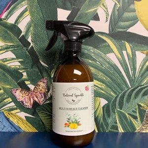 May include: A brown glass bottle with a black spray nozzle filled with a clear liquid. The bottle has a white label with the text "Natural Sparkle" and "Multi Surface Cleaner" in black lettering. The label also features a small illustration of lemons and flowers.