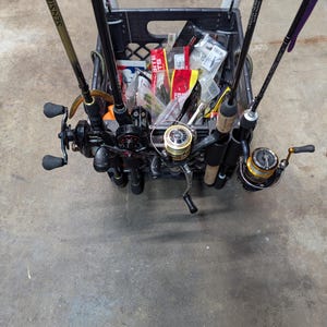 May include: A black plastic milk crate filled with fishing rods and tackle. Several fishing rods are secured in the crate, with reels attached. The crate contains various packages of fishing lures and other equipment, ready for a fishing trip.