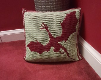 May include: A green crocheted pillow with a red dragon silhouette design. The pillow is sitting on a red carpet.