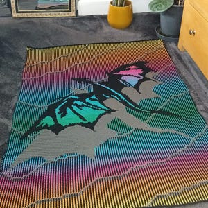 Puede incluir: Una colorida manta de punto con un diseño de dragón estilizado. La manta muestra un degradado de colores, incluyendo amarillo, naranja, rosa, azul y verde, con una silueta de dragón negro. La manta es rectangular y parece estar hecha de hilo suave.