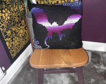 May include: A black and purple crocheted pillow with a white bat design. The pillow is sitting on a wooden chair with a dark brown finish.