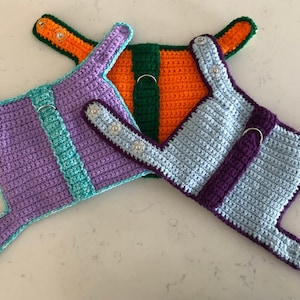 Pode incluir: Três arnês de cão de crochê em diferentes cores. O primeiro arnês é roxo e azul-turquesa, o segundo é laranja e verde, e o terceiro é azul claro e roxo.