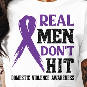 May include: White t-shirt featuring the text "REAL MEN DON'T HIT" in black, with a purple ribbon graphic. The words "DOMESTIC VIOLENCE AWARENESS" are printed below the main text, promoting awareness.