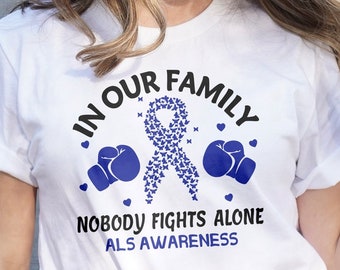 Op de afbeelding: Wit T-shirt met de tekst "IN OUR FAMILY NOBODY FIGHTS ALONE ALS AWARENESS". Het ontwerp bevat een blauw lint gemaakt van vlinders, blauwe bokshandschoenen en harten.