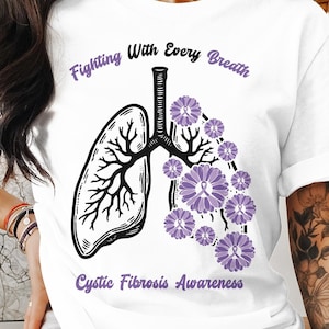 Puede incluir: Camiseta blanca con una cinta morada y el texto "Fighting With Every Breath Cystic Fibrosis Awareness" impreso en ella. El diseño presenta una imagen estilizada de pulmones con flores moradas.
