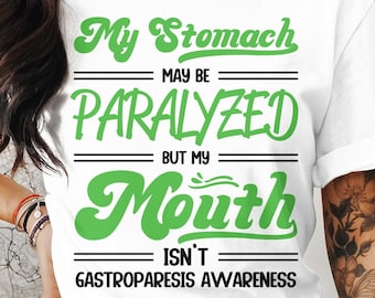May include: White t-shirt with green text that reads "My Stomach May Be Paralyzed But My Mouth Isn't Gastroparesis Awareness". The text is in a playful font. The shirt is a crew neck style.