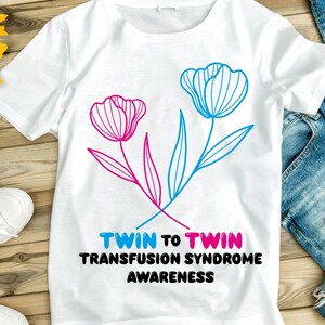 以下が含まれることがあります： ピンクとブルーの花柄がプリントされた白いTシャツに、「Twin to Twin Transfusion Syndrome Awareness」の文字が書かれています。