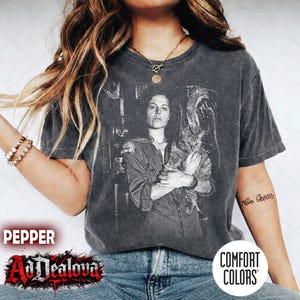 Peut inclure: T-shirt gris foncé avec un imprimé noir et blanc d'une femme tenant un chat. Le graphisme est tiré d'un film de science-fiction. Le t-shirt porte les inscriptions "Comfort Colors" et "AdDealova".