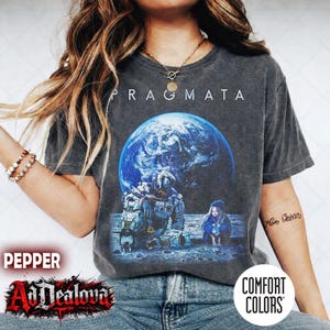 May include: Dark gray t-shirt with the word "PRAGMATA" above a graphic of Earth and two figures in a lunar setting. The shirt features a Comfort Colors tag and the AdDealova logo.