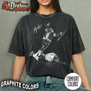 May include: Graphite-colored t-shirt featuring a black and white image of a hockey player in action, with a signature and the text "Graphite Colors". The shirt is from Comfort Colors.
