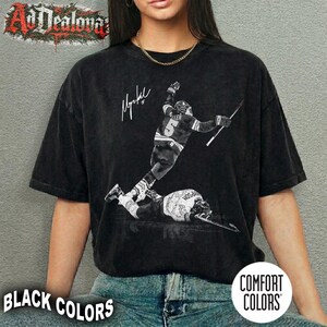 May include: Black Comfort Colors t-shirt featuring a black and white image of a hockey player in action, with a signature and the number 5. The shirt also has the text "BLACK COLORS" and "COMFORT COLORS".