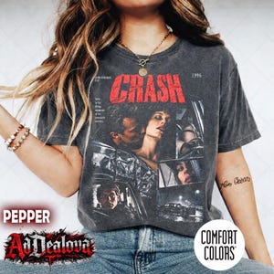 Könnte beinhalten: Ein anthrazitfarbenes Kurzarm-T-Shirt mit einem Grafikdruck im Vintage-Stil des Films Crash aus dem Jahr 1996. Das Design enthält den roten Schriftzug CRASH, mehrere schwarz-weiße Filmszenen und kleinen Text zur Filmproduktion.