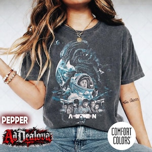 May include: Dark grey t-shirt featuring a graphic from the movie "Alien". The design includes an astronaut, an alien creature, and the word "Alien" in white. The shirt is a Comfort Colors brand.