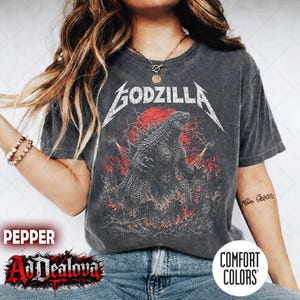 Op de afbeelding: Een antracietgrijs t-shirt met korte mouwen en een afbeelding van Godzilla in een stad met rode bliksem en een rode maan. Het woord GODZILLA staat in een wit, verweerd lettertype boven het wezen. Het shirt heeft een vintage wassing.